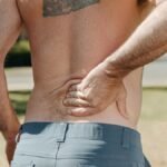 Shirtless man holding his lower back outdoors indicating pain or discomfort.