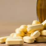 A close-up image of vitamin tablets spilling out of a transparent brown bottle on a wooden surface.