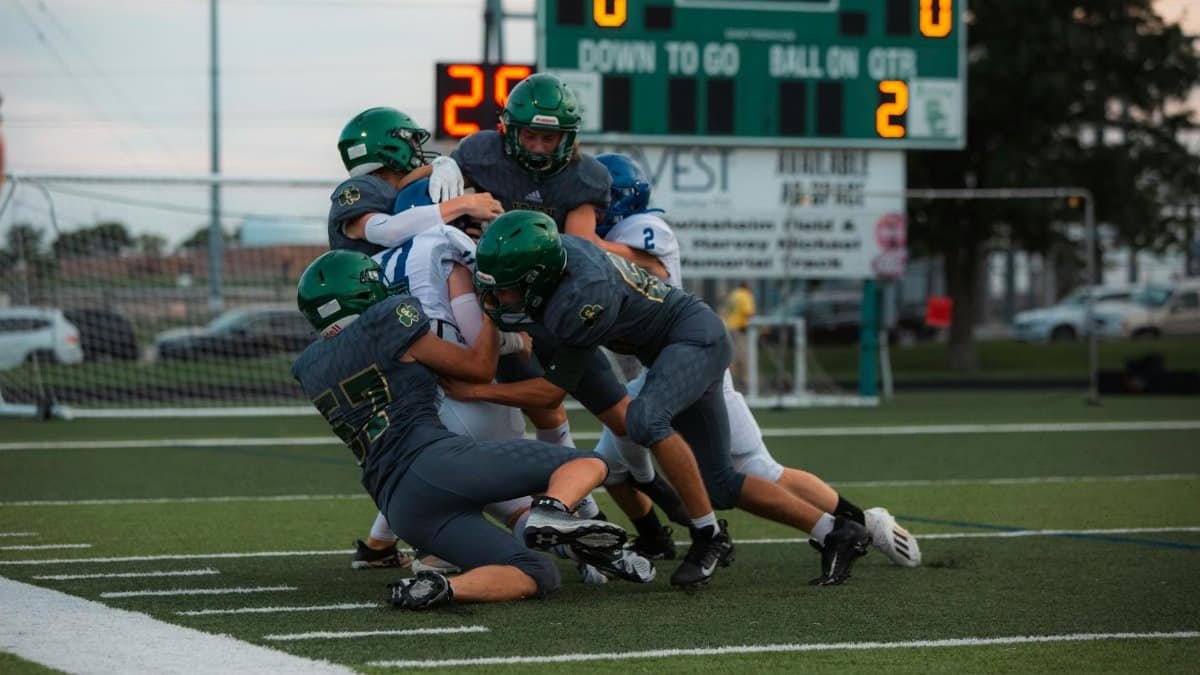 Football players engaged in an intense tackle during a high school game on a green field.