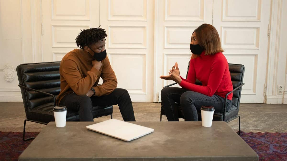 Focused African American colleagues in protective masks looking at each other while having conversation about work at table with laptop and paper cups of takeaway coffee