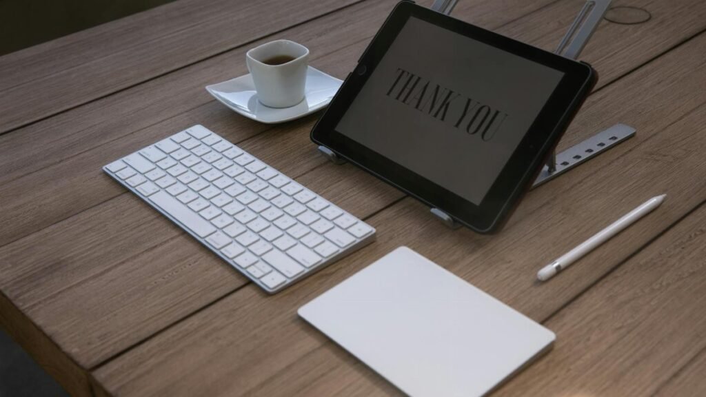 A contemporary workspace features a tablet displaying 'Thank You', a keyboard, an Apple pencil, and a coffee cup on a wooden desk.