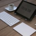 A contemporary workspace features a tablet displaying 'Thank You', a keyboard, an Apple pencil, and a coffee cup on a wooden desk.
