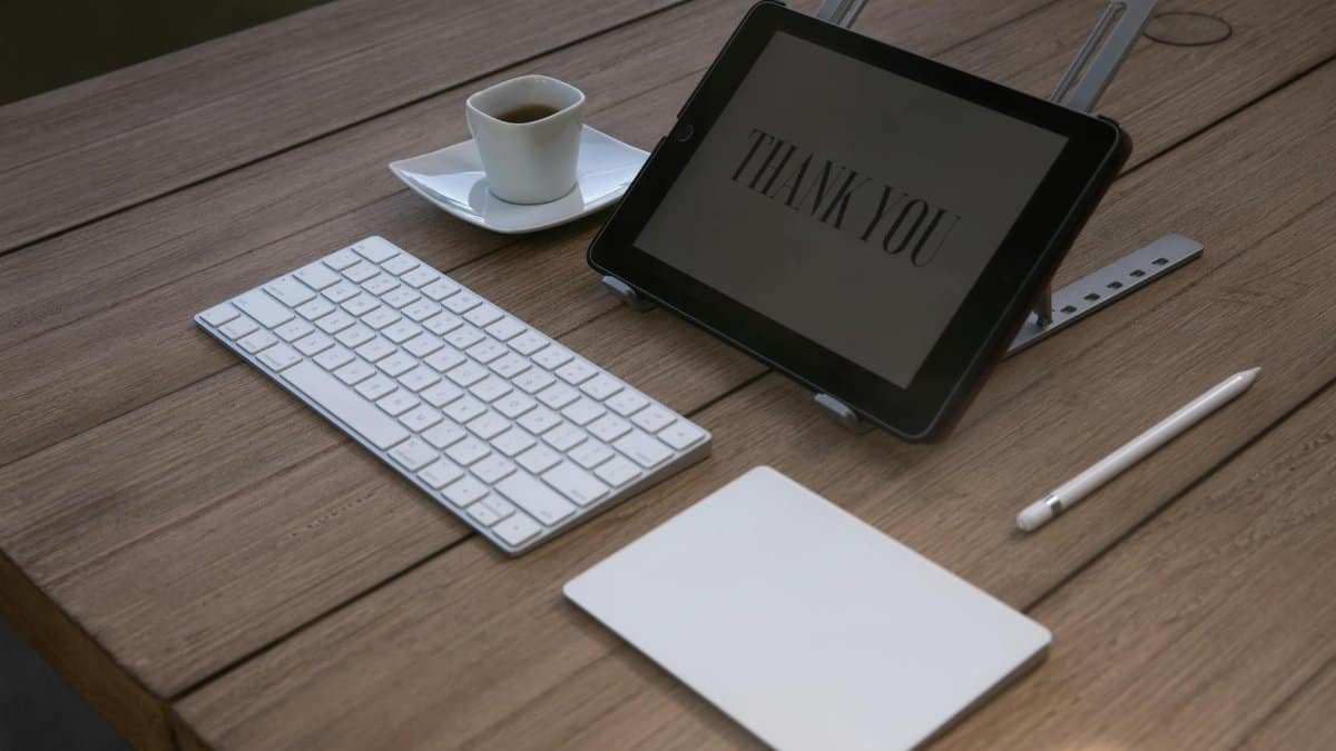 A contemporary workspace features a tablet displaying 'Thank You', a keyboard, an Apple pencil, and a coffee cup on a wooden desk.