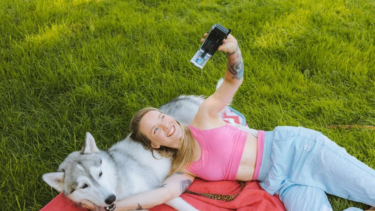 A cheerful woman in a pink tank top takes a selfie with her Siberian Husky while lying on a red blanket in a grassy park.