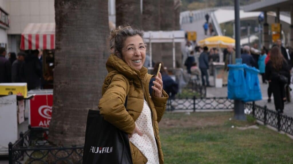 Smiling woman with phone and Fujifilm bag at bustling outdoor market in İzmir, Türkiye.