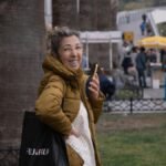 Smiling woman with phone and Fujifilm bag at bustling outdoor market in İzmir, Türkiye.