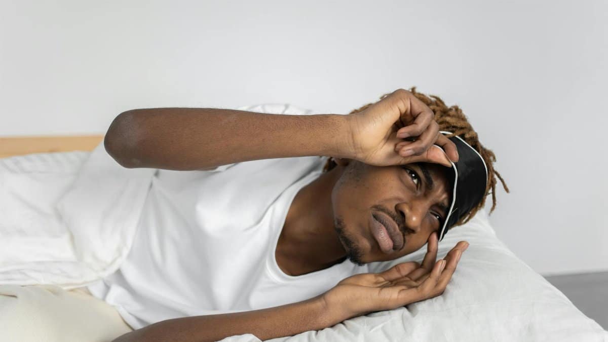 Young man removes sleep mask, starting his day in bed, indoors.