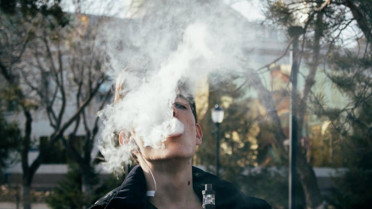 Portrait of a young man vaping in a park, releasing clouds of vapor on a clear day.