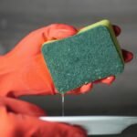 Close-up of hands in rubber gloves using a sponge to wash a ceramic plate over a sink.