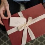 Close-up of hands holding a red gift box with a white ribbon and a message tag.