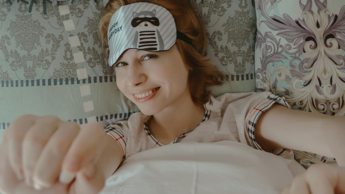 A cheerful woman in pajamas stretches and smiles in bed, wearing a cute sleep mask.