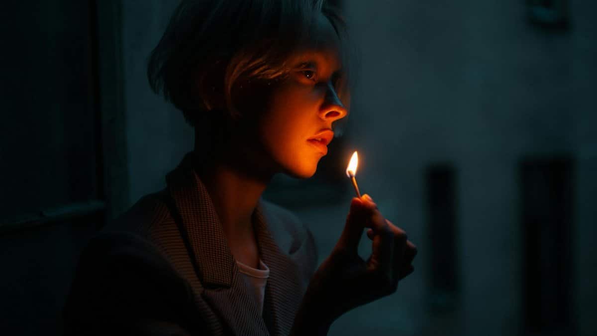 A contemplative woman holds a lit matchstick, casting warm light on her face in the dark.