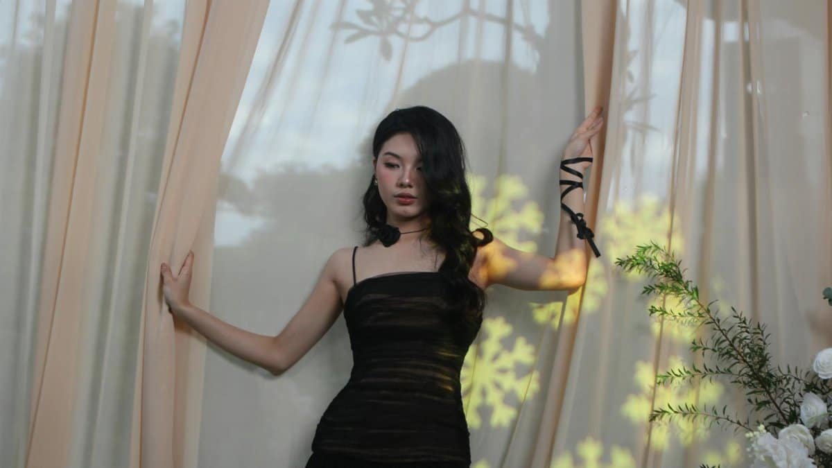 A stylish woman poses by sheer curtains with floral shadows in an elegant indoor setting.