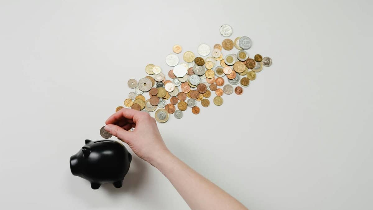 Person placing coin into a black piggy bank with scattered coins, symbolizing savings and finance.