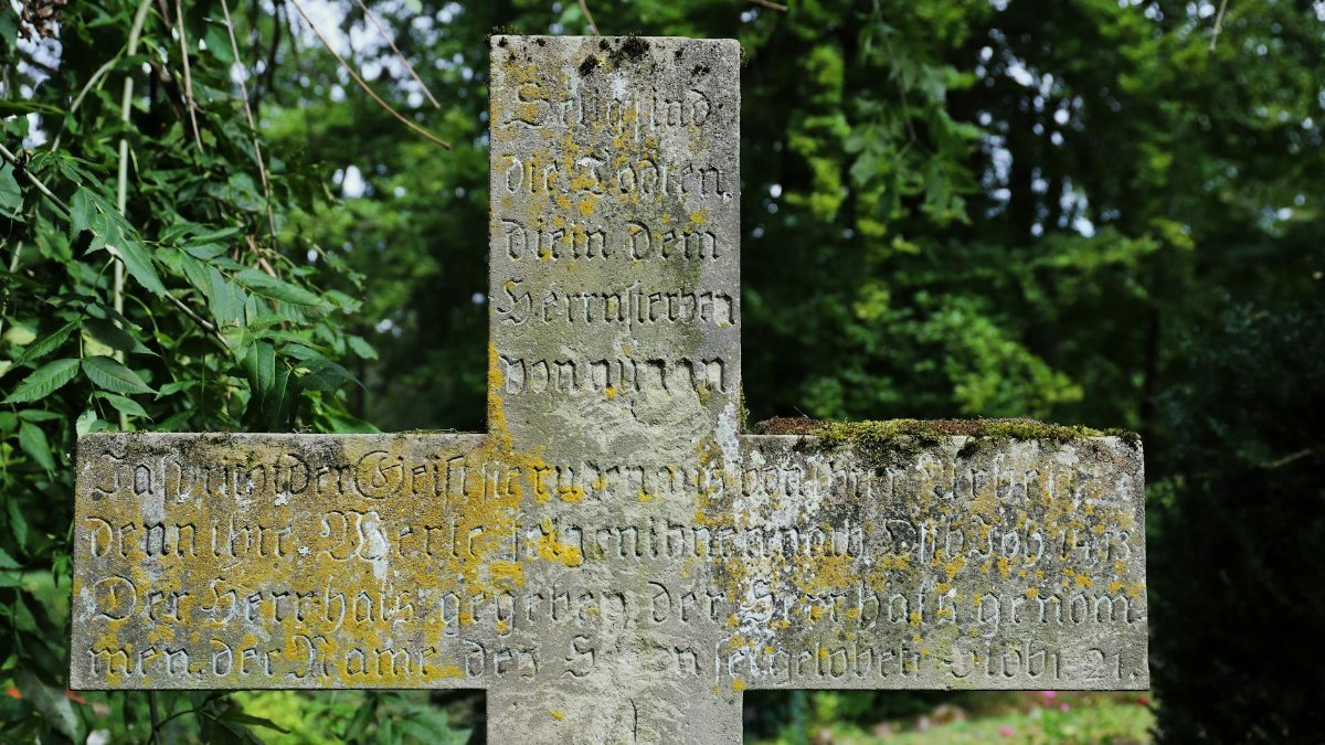 Weathered stone cross with engraved text in a German cemetery surrounded by greenery.