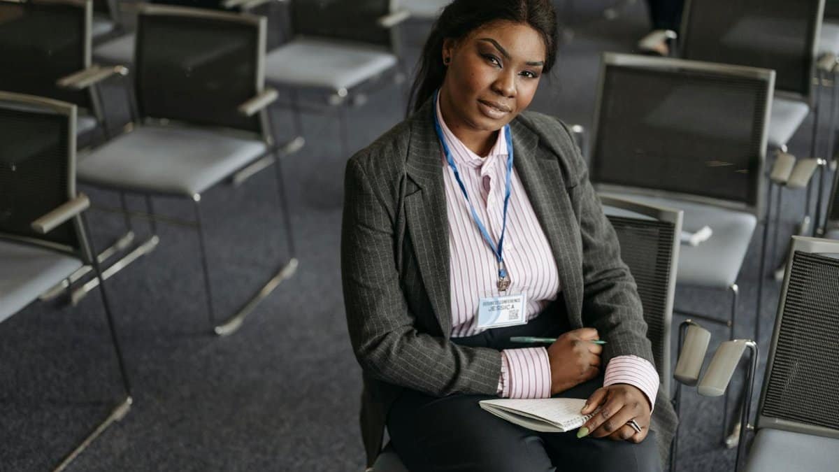 Confident woman in business attire sitting in a conference room with notepad and ID badge.