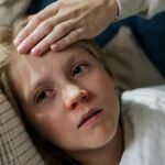 Young child lying down with a hand checking for fever, indicating illness or flu symptoms.