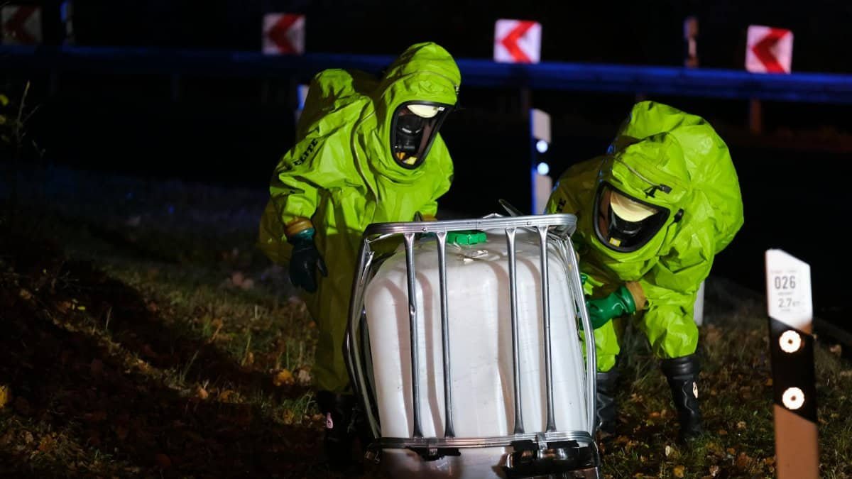 Two firefighters in hazmat suits managing a chemical spill by the roadside at night.