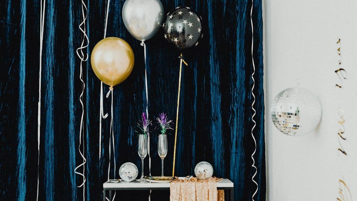 Stylish New Year Eve party decor featuring balloons, disco balls, and wineglasses against a blue curtain.