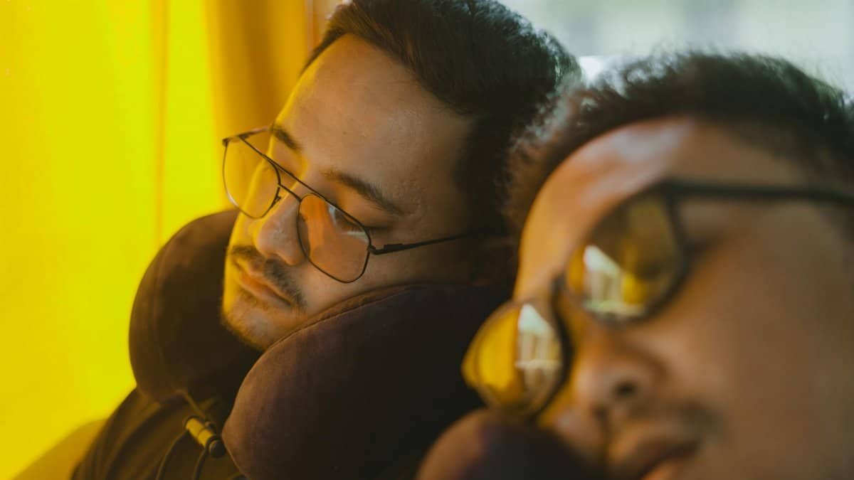 Two men wearing eyeglasses sleep on a bus, using neck pillows for comfort during the journey.