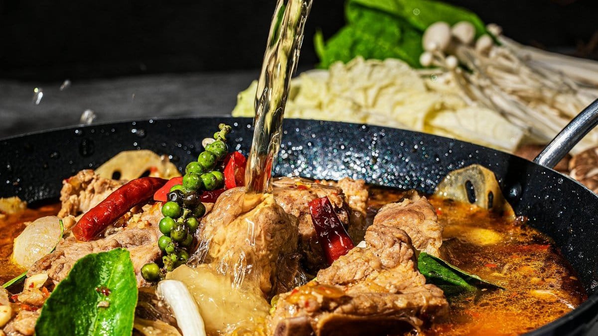 A rich and spicy hot pot with meat, vegetables, and spices pouring in a simmering broth.