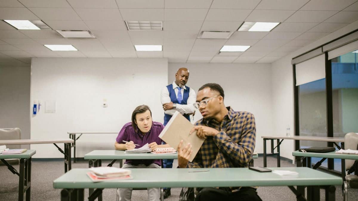 Students in a classroom secretly talking while a teacher observes from behind.