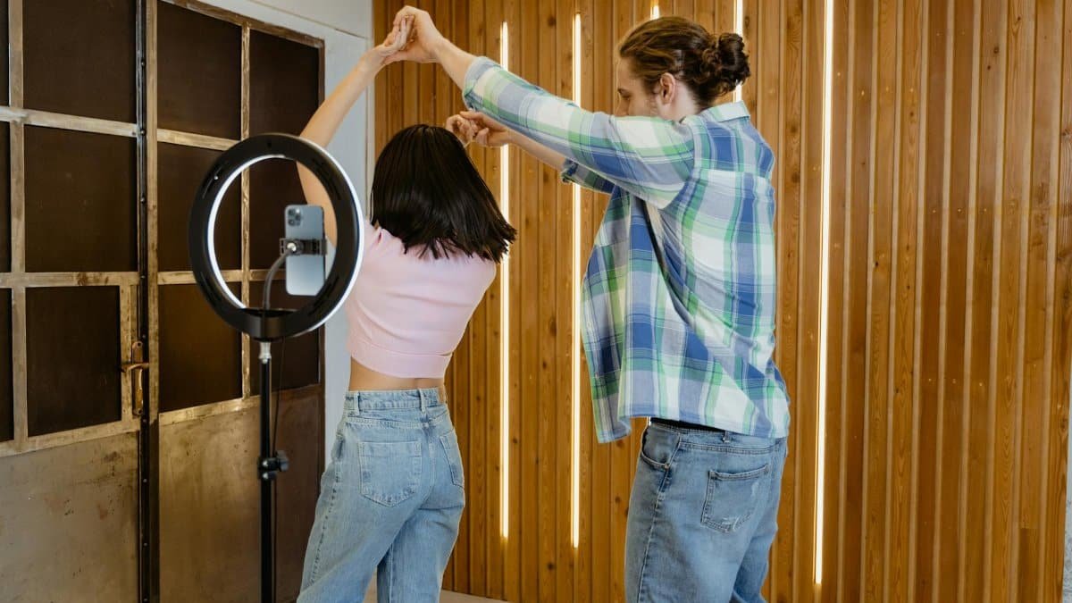 A young couple dances indoors with a ring light recording them, capturing a lively moment.