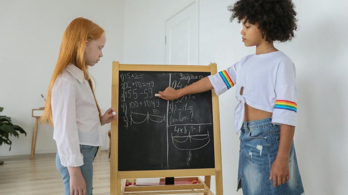 Two girls solving math equations on a chalkboard in a bright classroom setting.