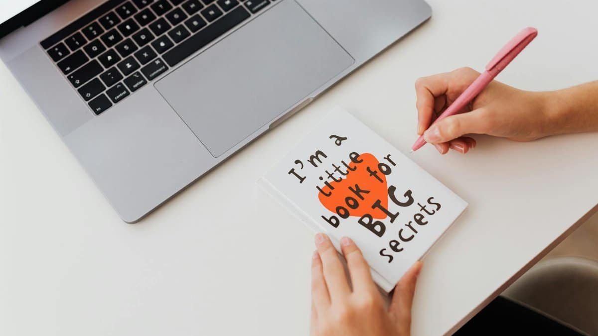 Overhead view of a woman writing in a notebook labeled 'I'm a little book for BIG secrets' beside a laptop.