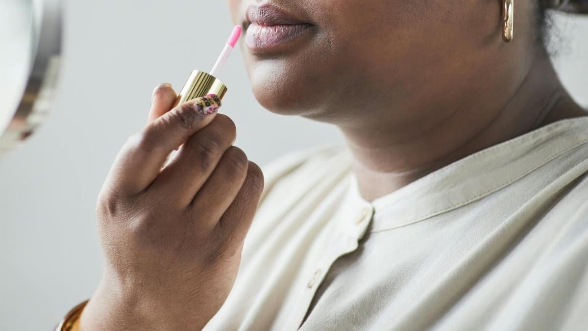 A young woman applies lip gloss in a bright setting, highlighting natural beauty and skincare routines.