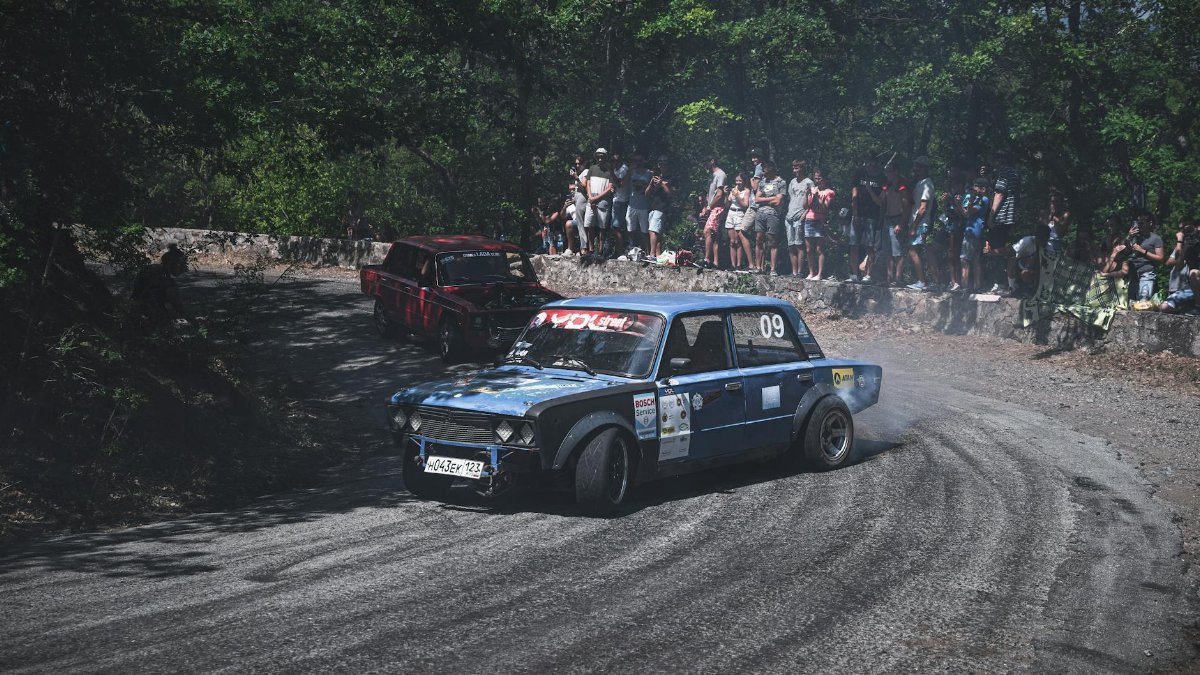 A rally car drifts around a tight turn on a forested track as spectators watch.