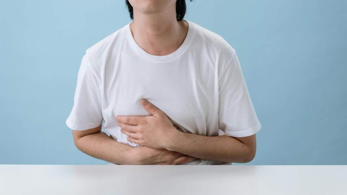 An adult man in a white shirt holds his stomach, indicating pain and discomfort, with a light blue background.