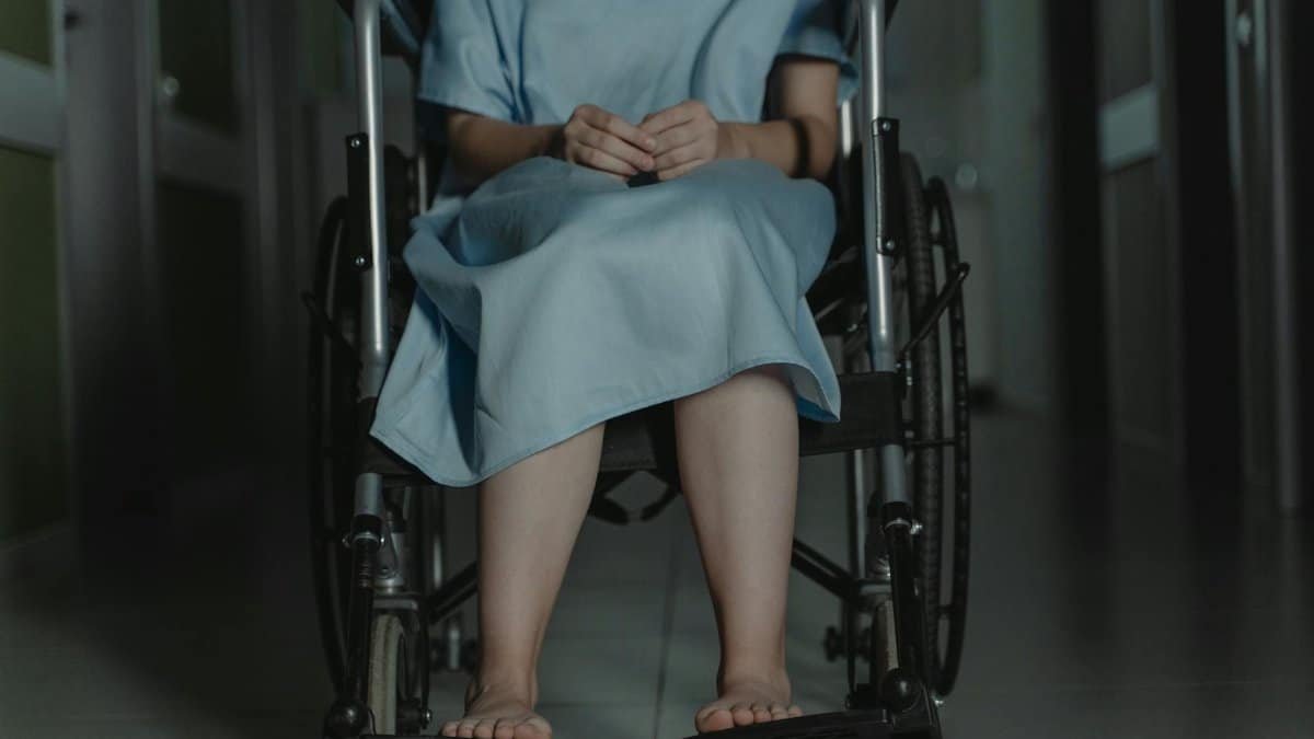 Person sitting barefoot in a wheelchair wearing a hospital gown in a dim hallway.