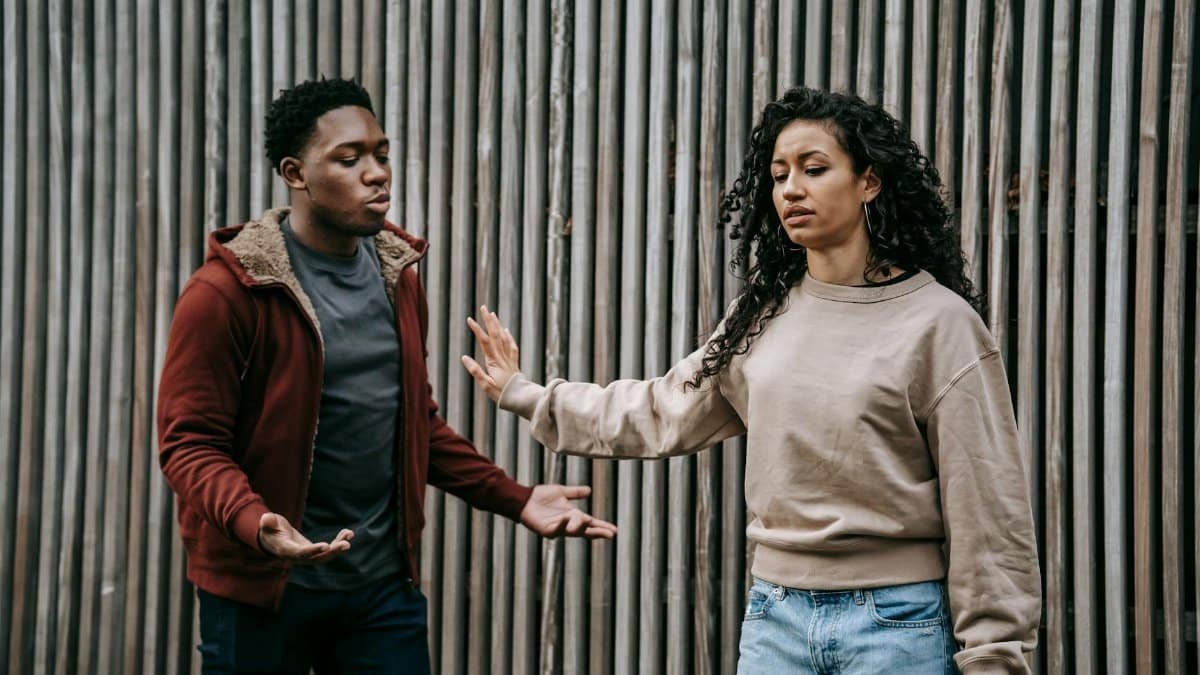 Insulted ethnic girlfriend with outstretched arm and irritated African American boyfriend having quarrel near wooden wall on street during breakup