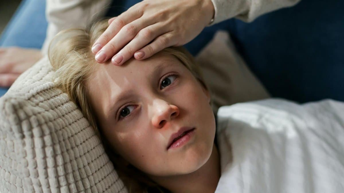 Young child lying down with a hand checking for fever, indicating illness or flu symptoms.