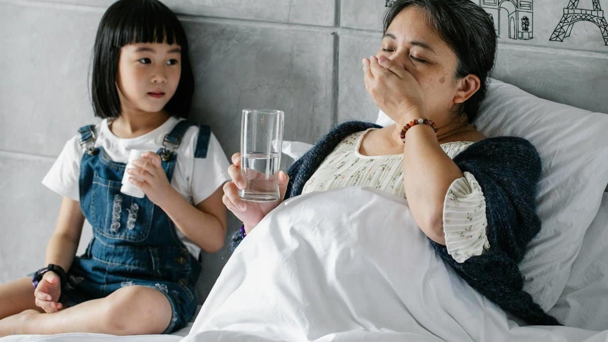 Cute Asian little girl sitting on soft bed and looking at sick grandmother drinking medicine on sunny day