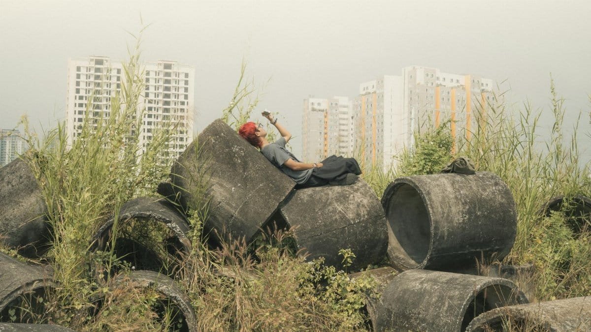 A man with red hair relaxes on large concrete pipes in an overgrown urban area with high-rise buildings in the background.