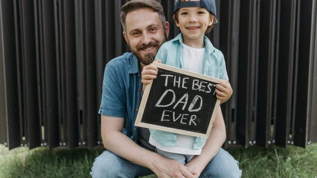 A father and son smiling together holding a 'Best Dad Ever' sign outside.
