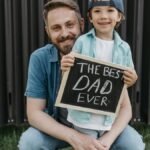 A father and son smiling together holding a 'Best Dad Ever' sign outside.