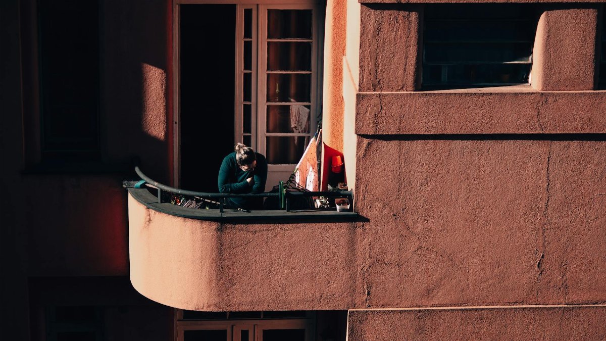 A person reflects quietly on a balcony amidst an urban scene, captured in deep shadows and warm tones.
