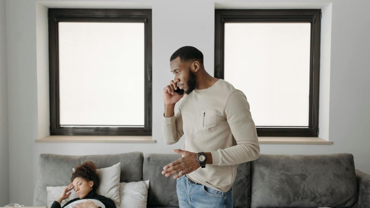 A concerned man makes a phone call while a woman sleeps unwell in bed, indicating urgency and care.