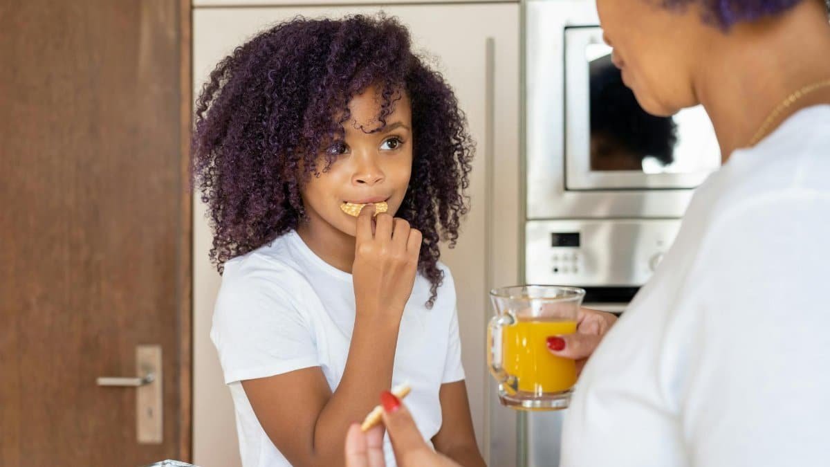 A girl eating a cracker while her mother holds a glass of orange juice in a kitchen setting.