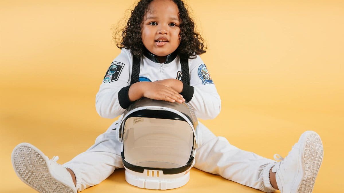 Cute child wearing a spacesuit with helmet, sitting on yellow backdrop.