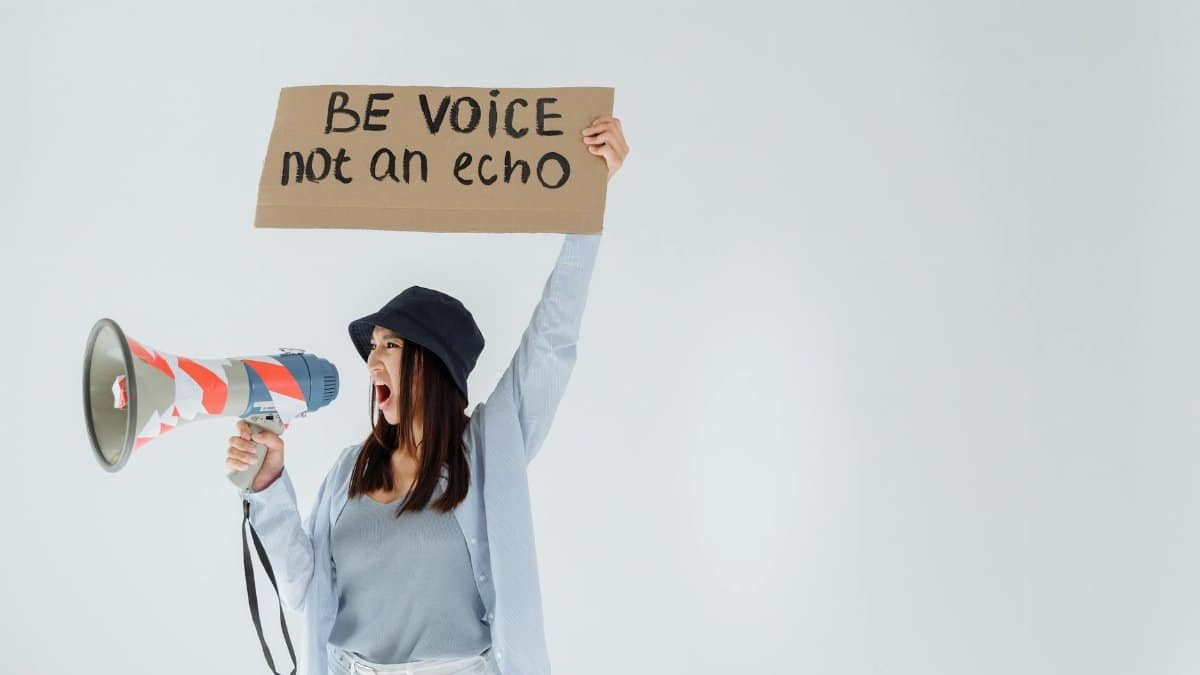 Empowered woman shouts message through megaphone, holding sign that reads 'Be voice, not an echo'.