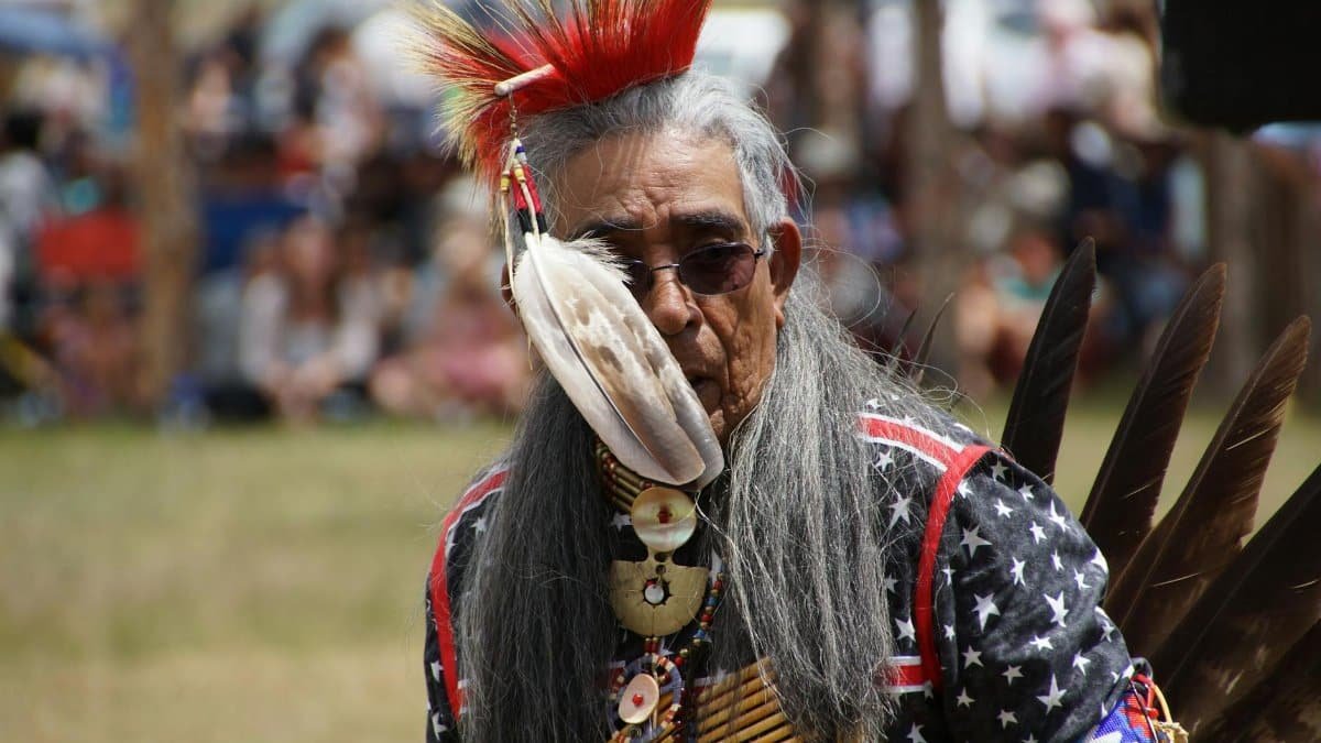Senior Native American man in elaborate headdress and attire at a cultural gathering.