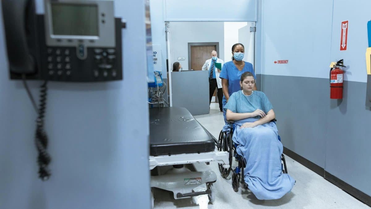 A nurse helps a patient in a wheelchair down a hospital corridor, reflecting care and medical professionalism.