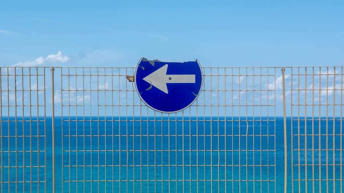 A blue arrow sign on a metal fence overlooks the calm ocean under a clear sky.