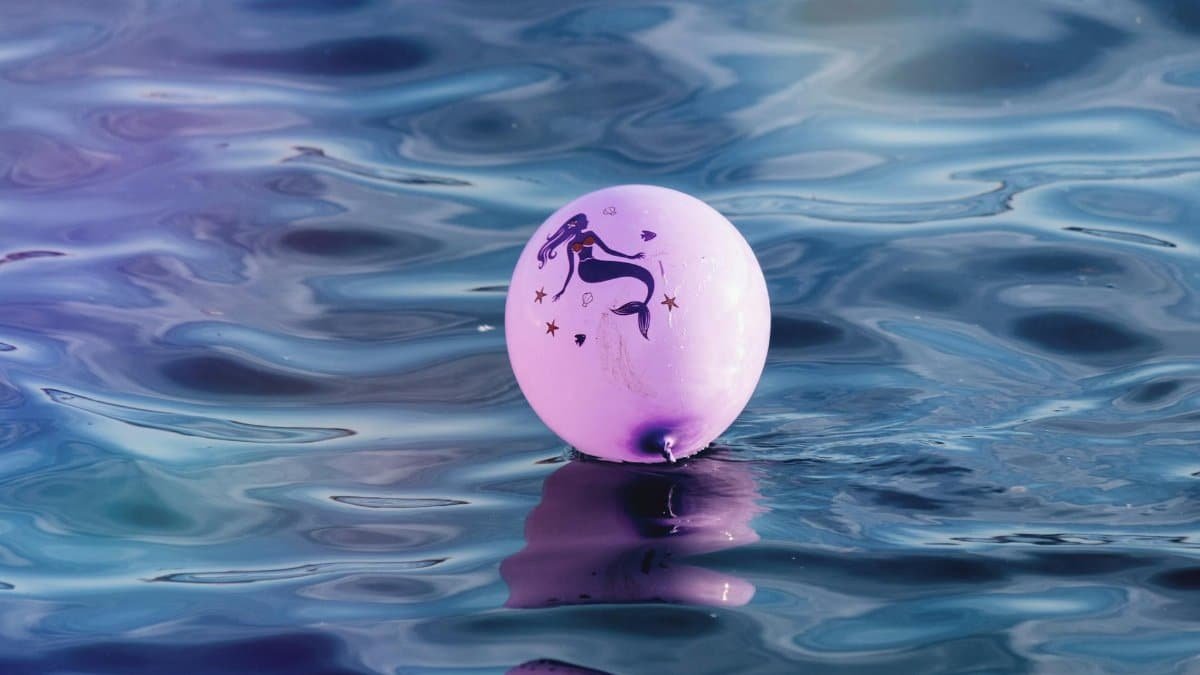 A pink balloon featuring a mermaid design floats on a calm, reflective water surface.