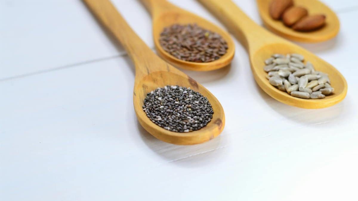 A variety of seeds and nuts in wooden spoons displayed on a white surface.