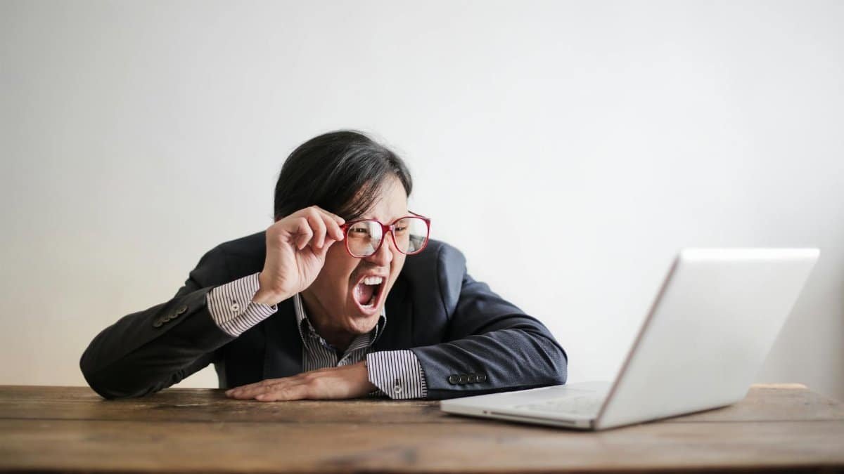 Modern Asian man in jacket and glasses looking at laptop and screaming with mouth wide opened on white background